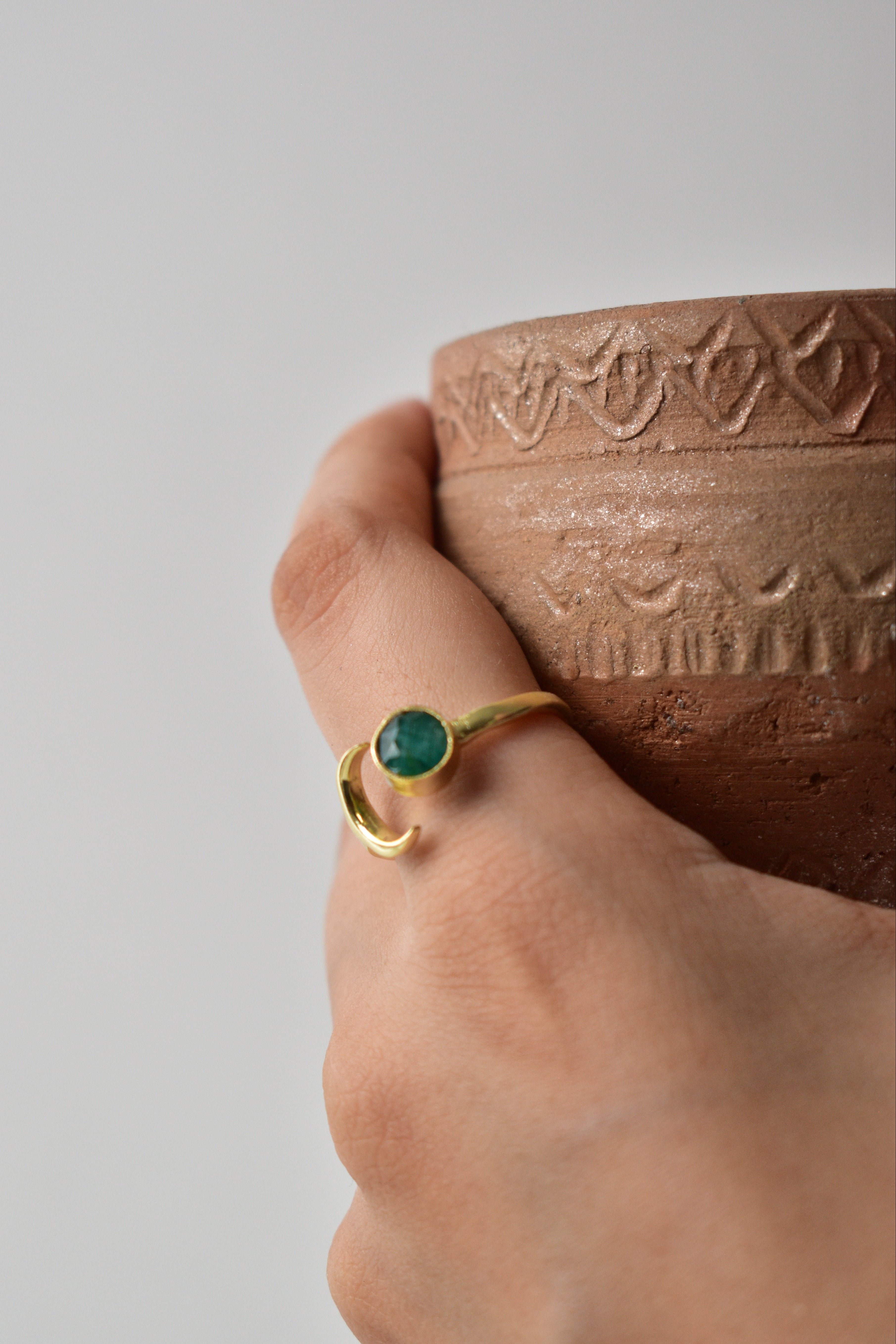 Hand wearing a gold ring with a green gemstone, holding a textured brown object against a gray background
