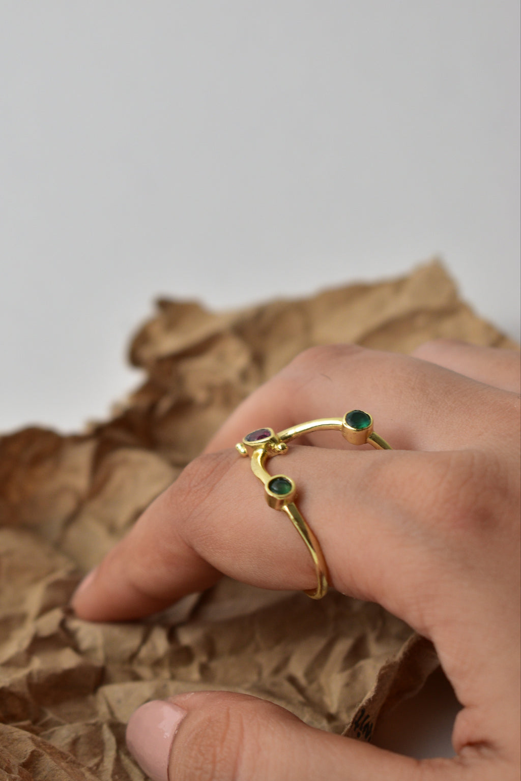 Gold ring with green gemstones on a finger, held against crumpled brown paper.
