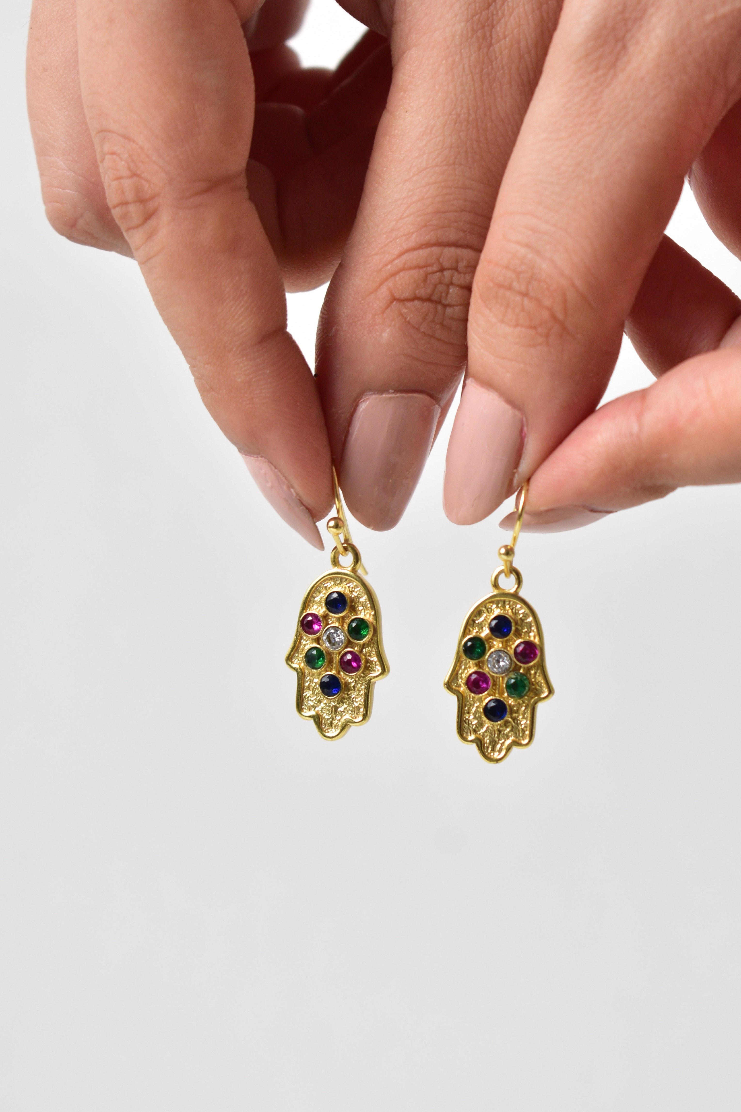 Gold earrings with colorful gemstones held by a hand against a light gray background