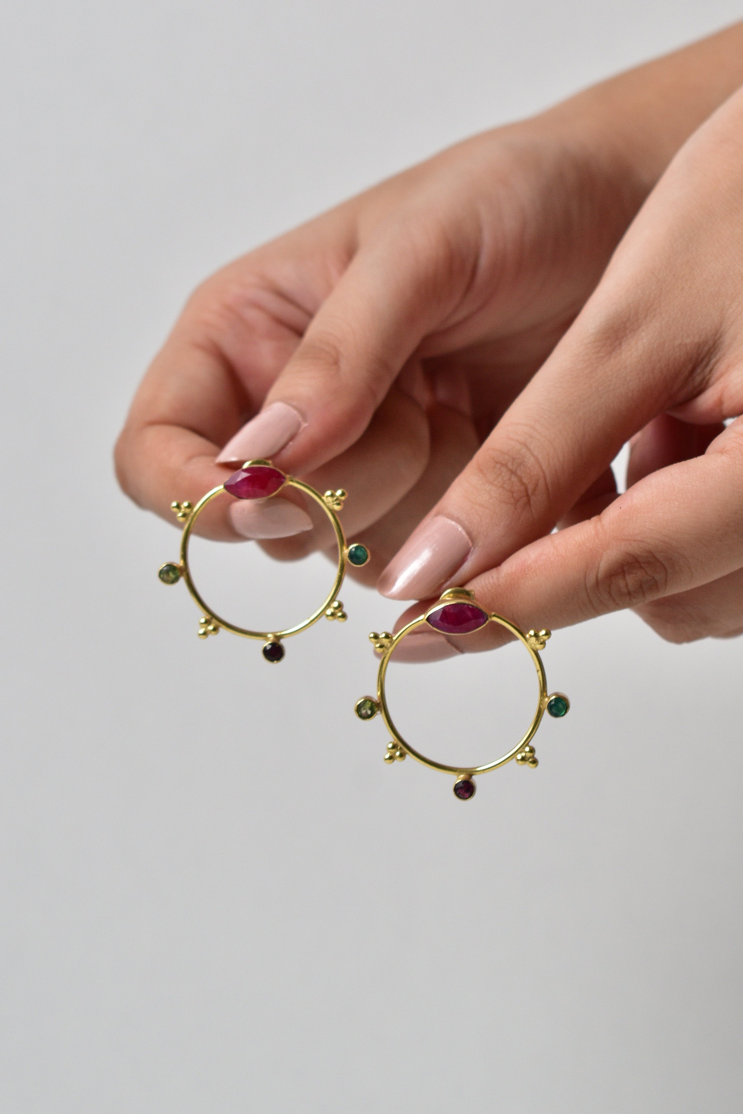 Gold hoop earrings with gemstones held by a hand against a plain background