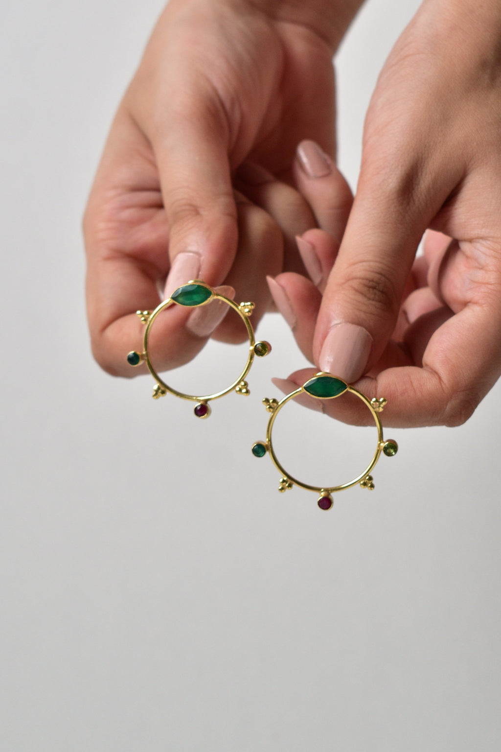 Gold hoop earrings with green gemstones held by a hand against a plain background