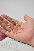 Gold earrings with green and red stones held in a hand against a blurred background