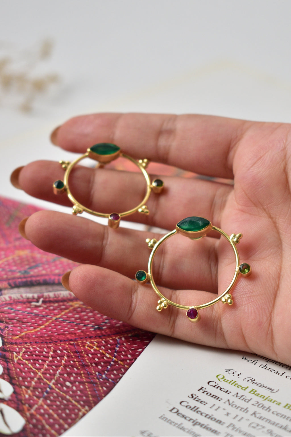 Gold hoop earrings with green gemstones held in a hand against a blurred background