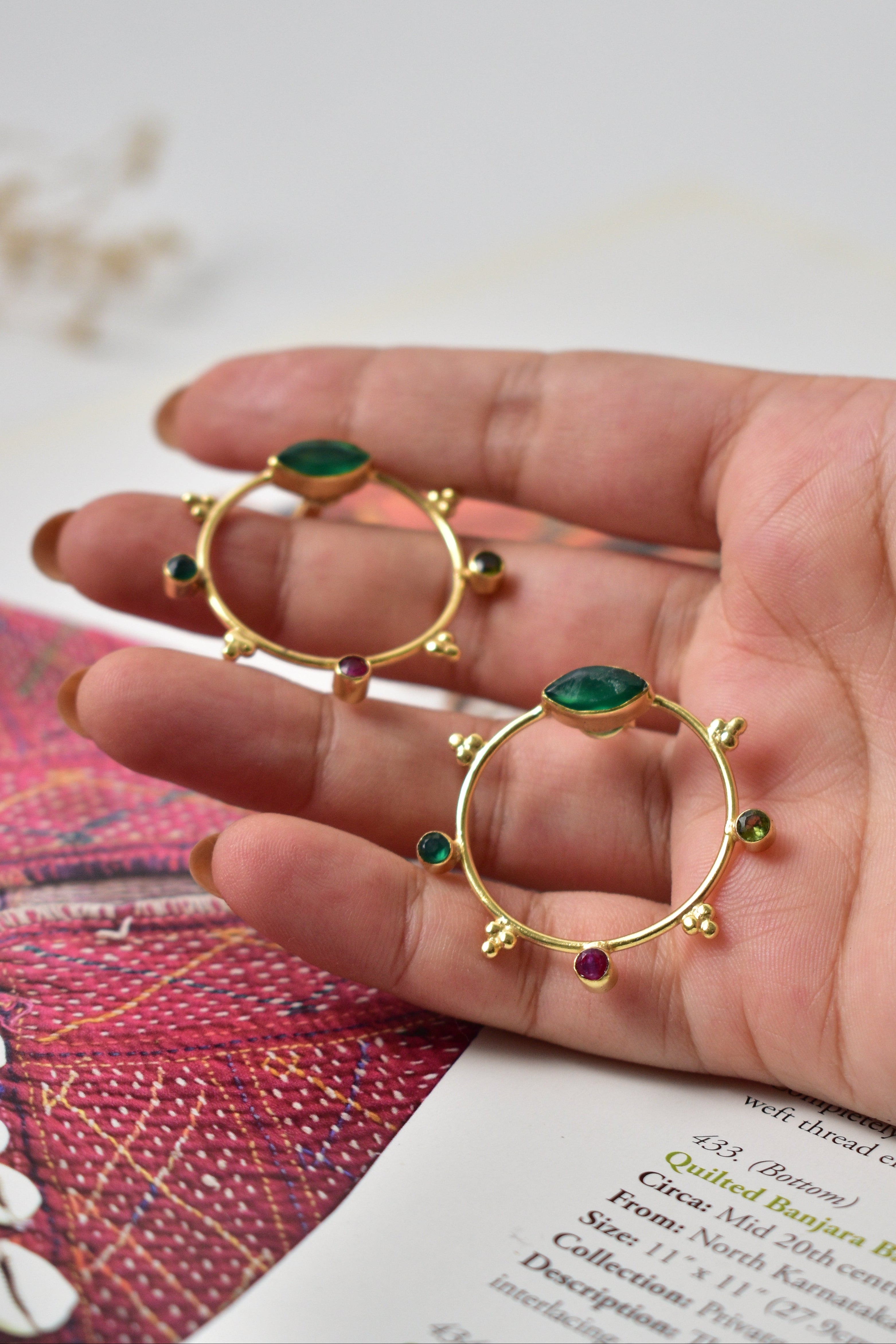 Gold hoop earrings with green gemstones held in a hand against a blurred background
